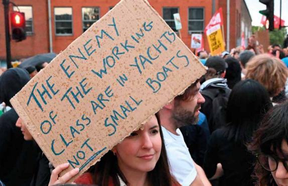 Woman holding a sign that reads "The enemy of the working class are in yachts not small boats"