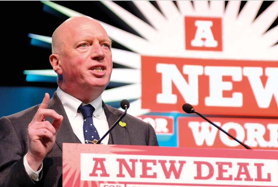 Matt Wrack speaking from podium in front of a new deal for working people sign