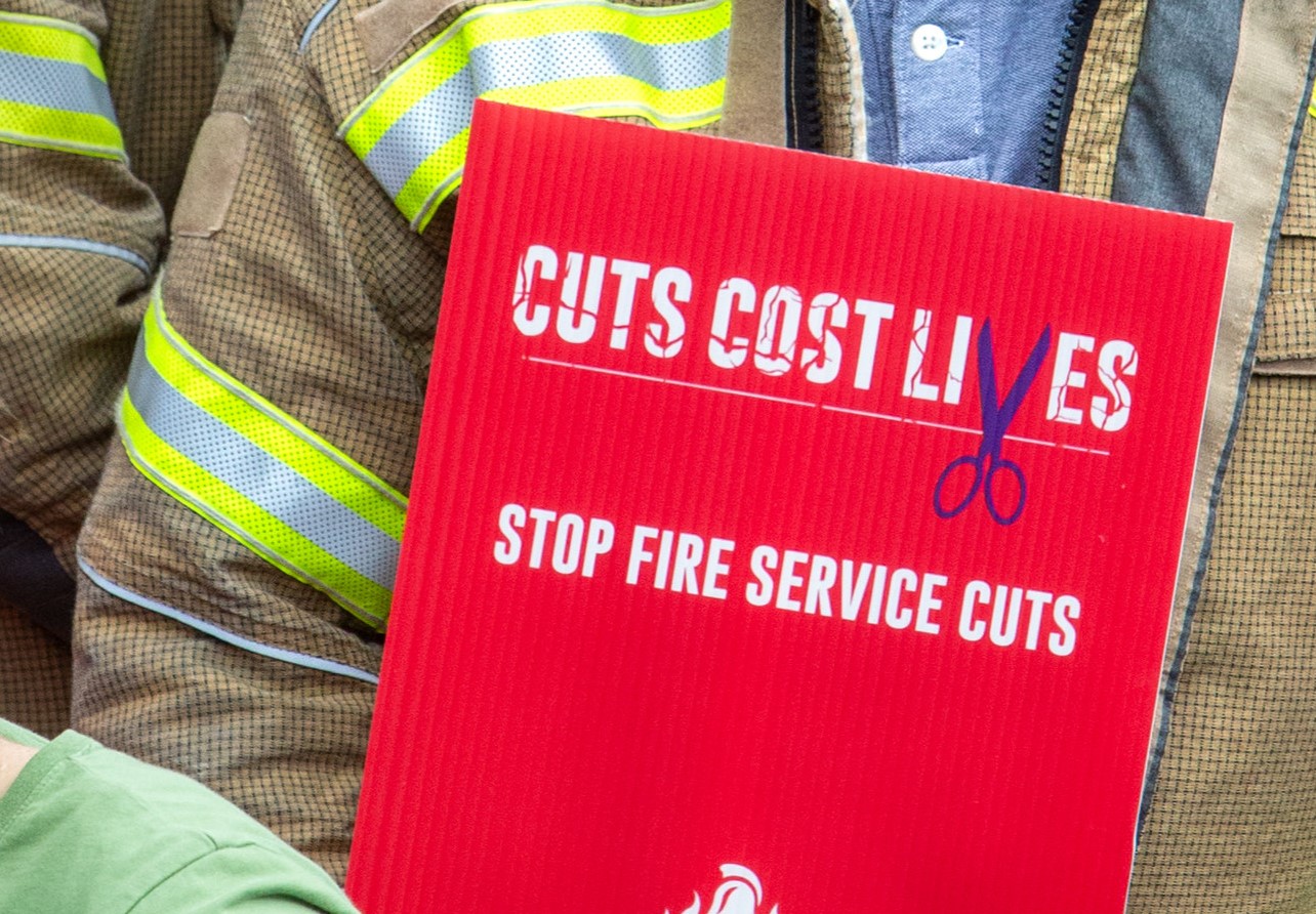 A firefighter holds a sign reading 'Cuts cost lives, stop fire service cuts'