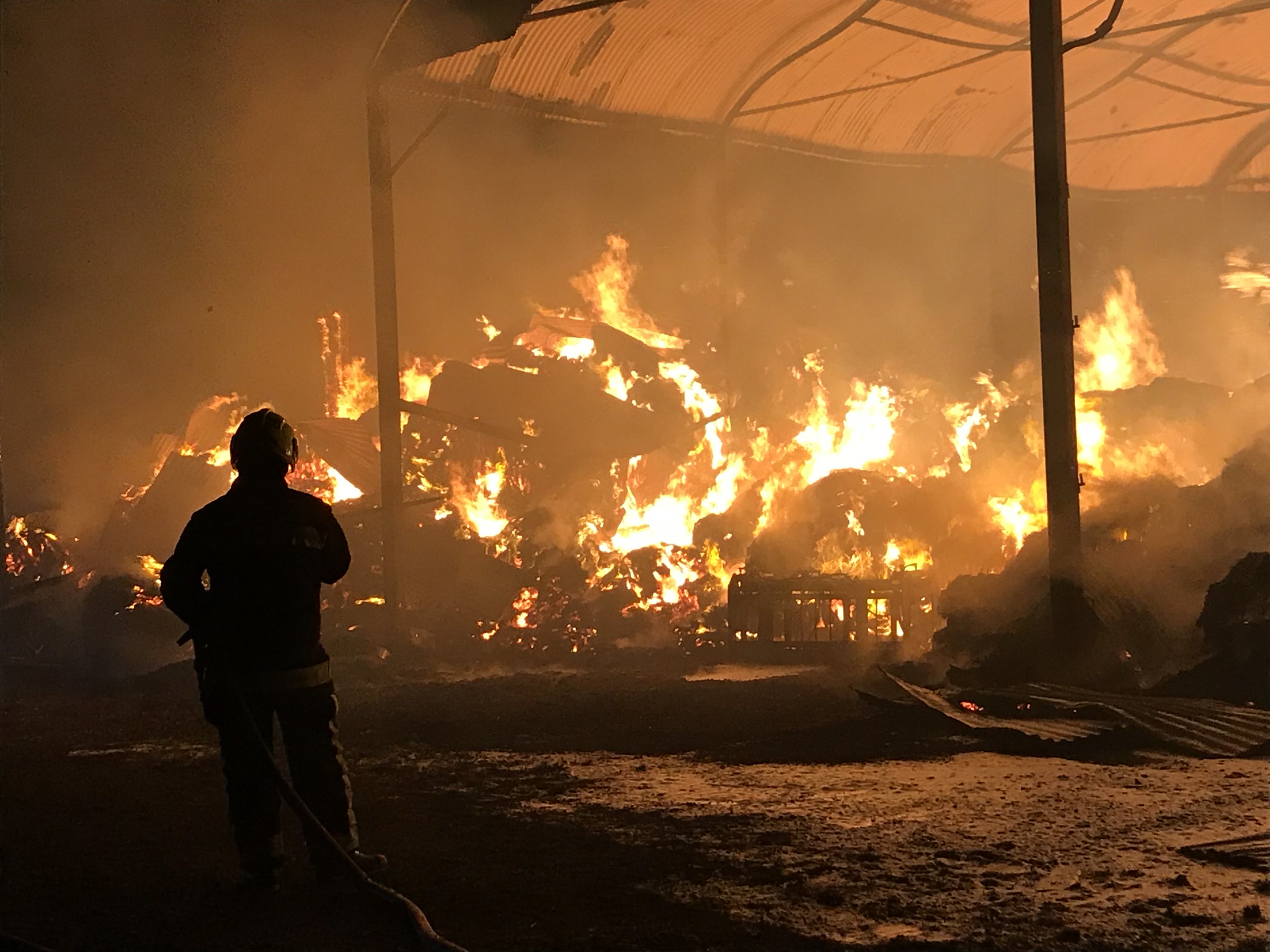 A picture of a barn fire, mostly orange, with one firefighter in the left foreground with their back turned