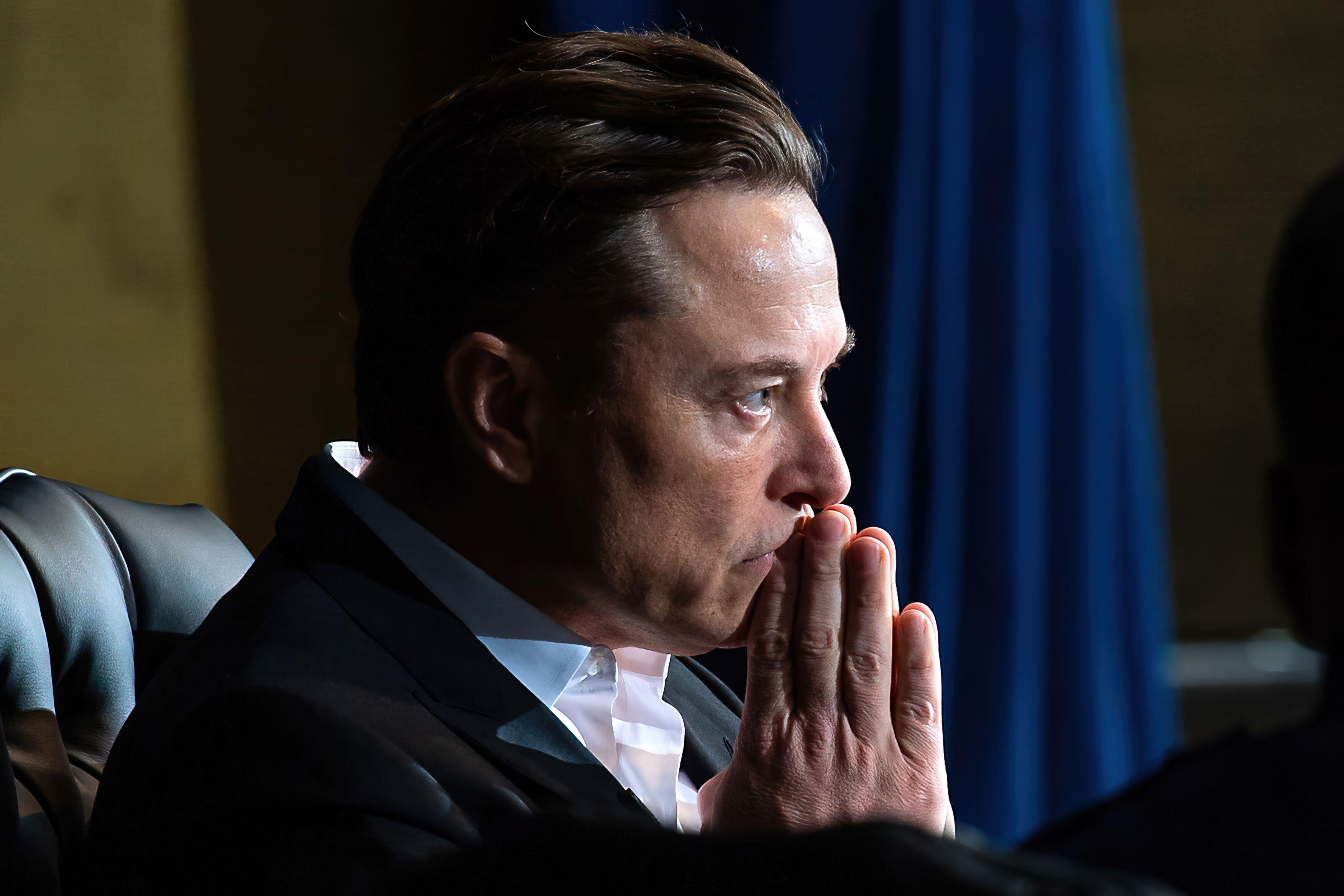 Elon Musk thinking and looking forwards with his hands up to his mouth
