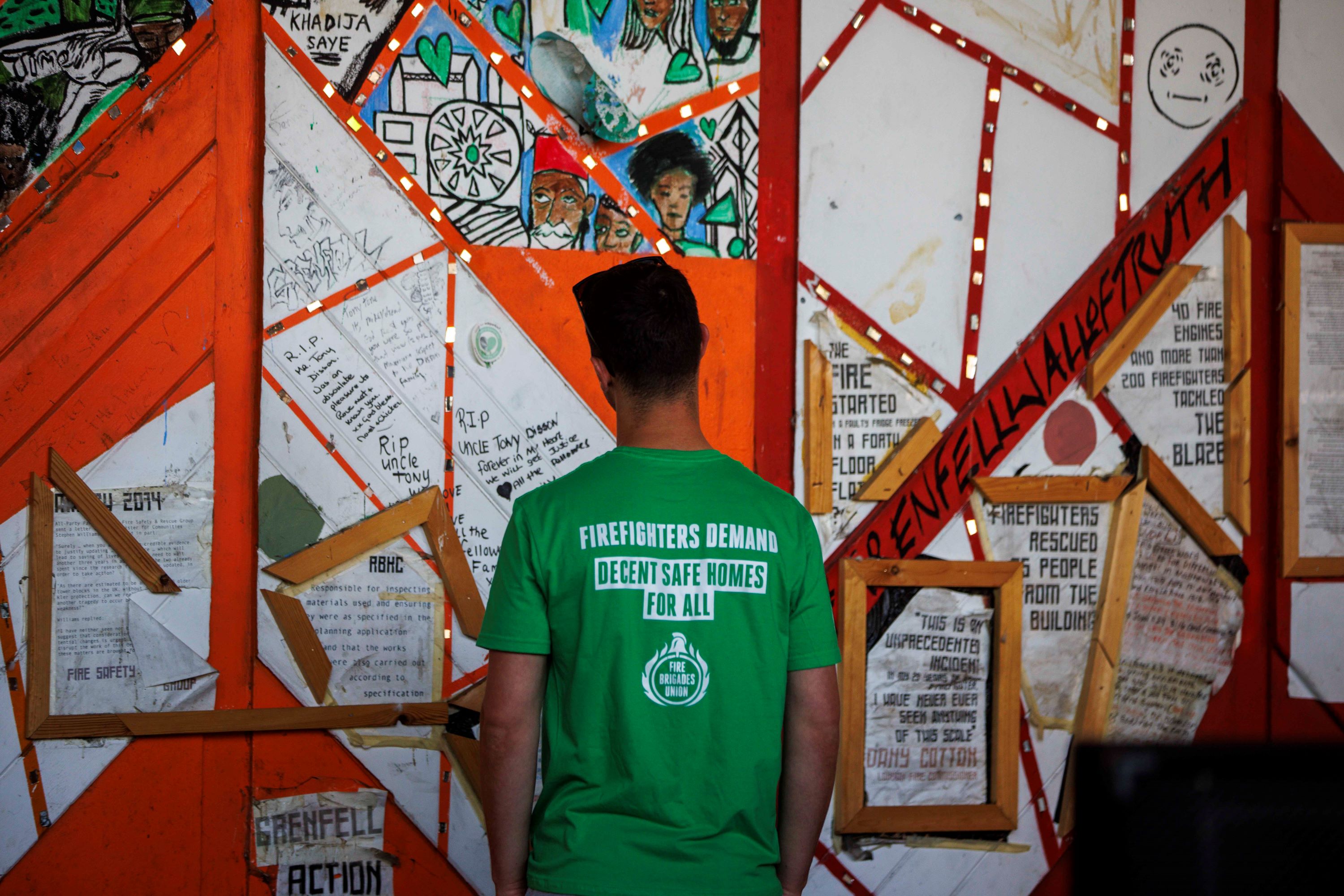 Firefighter wearing a t shirt reading 'FBU demands safe homes for all' looks at the Grenfell memorial wall