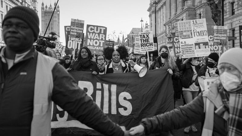 Black and white photo of a march outside Parliament with women holding signs saying cut warfare not welfare