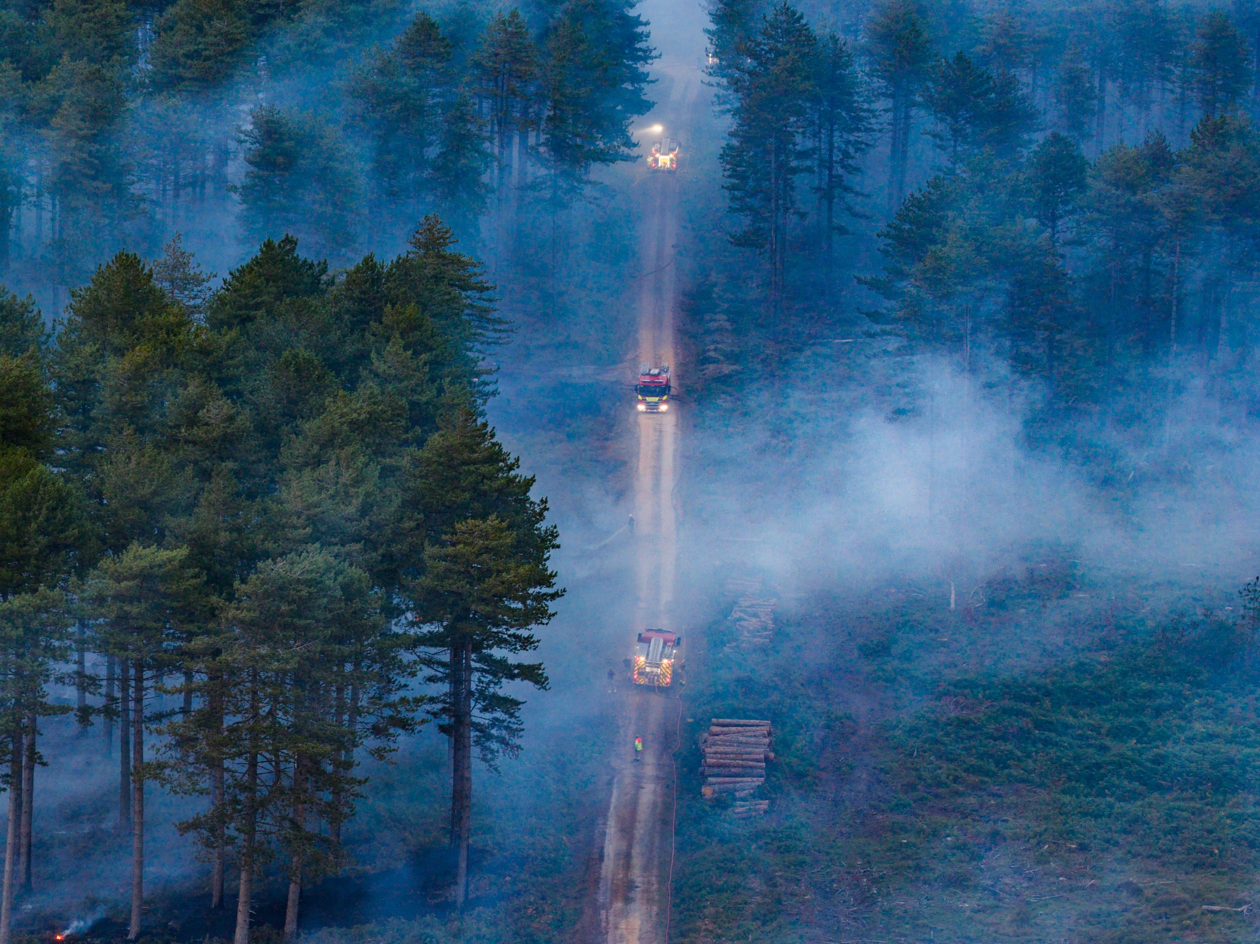 Aerial view of two fire appliances driving on a long road through smoke of wildfire