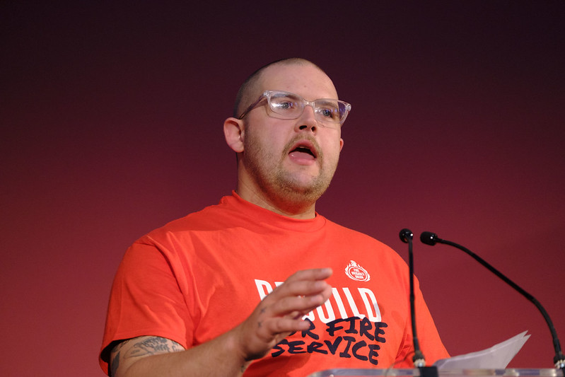 Lewis Clark speaking from the podium wearing a rebuild our fire service t shirt