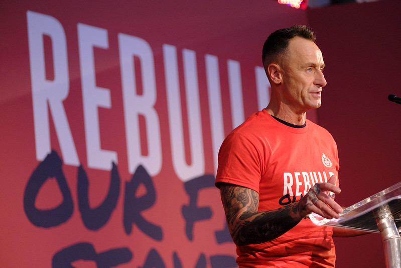Craig Elson speaks from a podium in front of a red backdrop with slogan rebuild our fire service in large lettering
