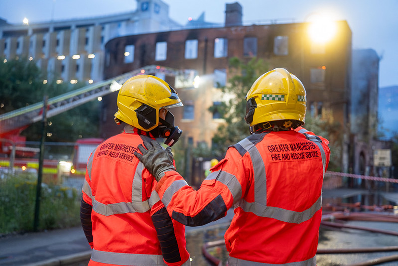 A firefighter puts their arm on the shoulder of a colleague looking at a burned out building