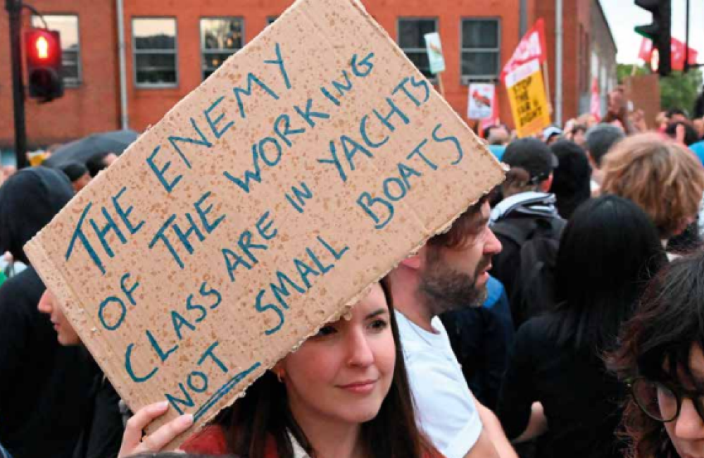 Woman holding a sign that reads "The enemy of the working class are in yachts not small boats"