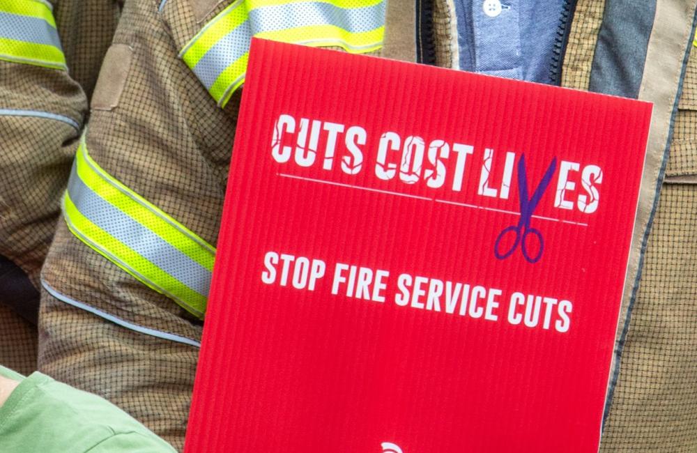 A firefighter holds a sign reading 'Cuts cost lives, stop fire service cuts'