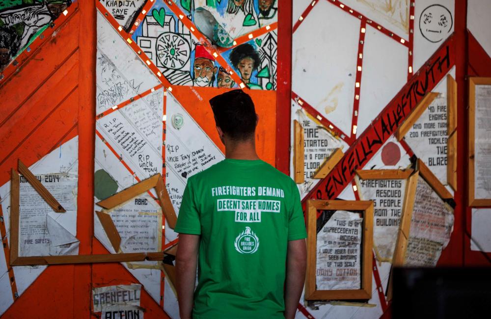 Firefighter wearing a t shirt reading 'FBU demands safe homes for all' looks at the Grenfell memorial wall