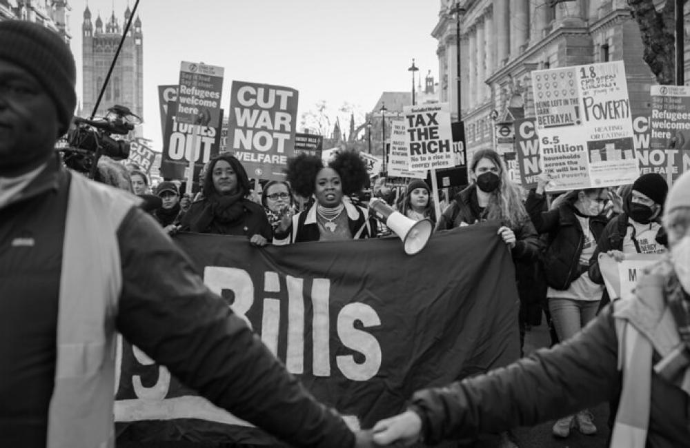 Black and white photo of a march outside Parliament with women holding signs saying cut warfare not welfare