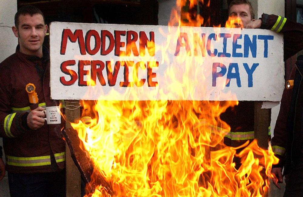 Firefighters behind a picket line brazier hold up a placard reading "modern service, ancient pay"