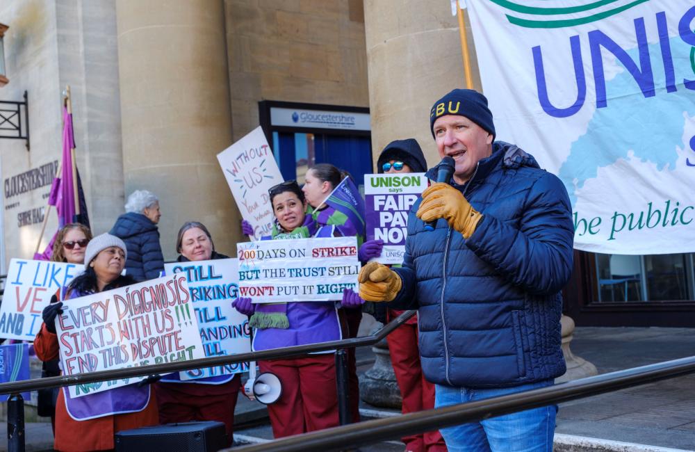 Tam Macfarlane wearing an FBU beanie speaks with a mic at a rally of striking phlebotomists