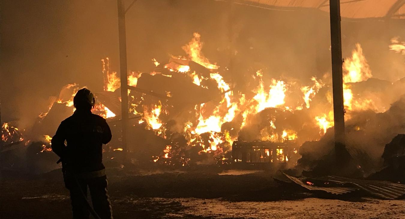 A picture of a barn fire, mostly orange, with one firefighter in the left foreground with their back turned