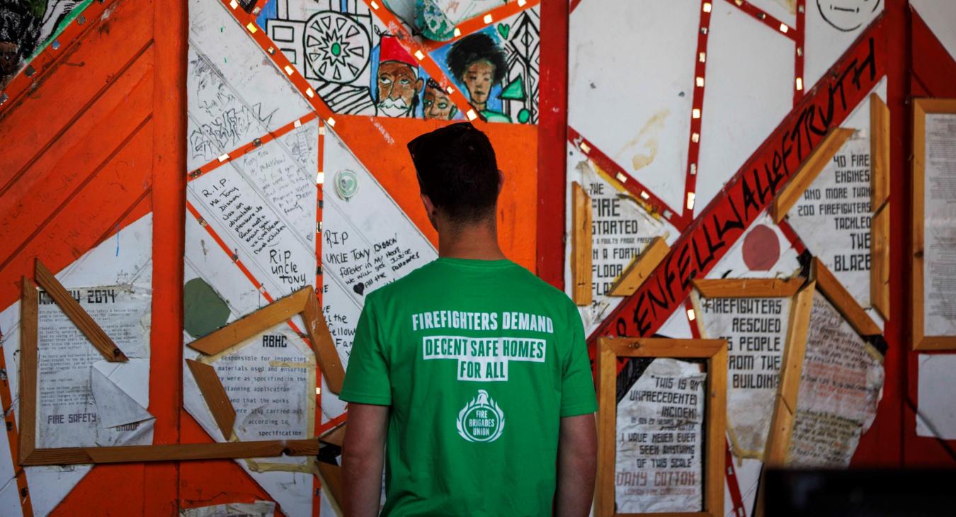 Firefighter wearing a t shirt reading 'FBU demands safe homes for all' looks at the Grenfell memorial wall