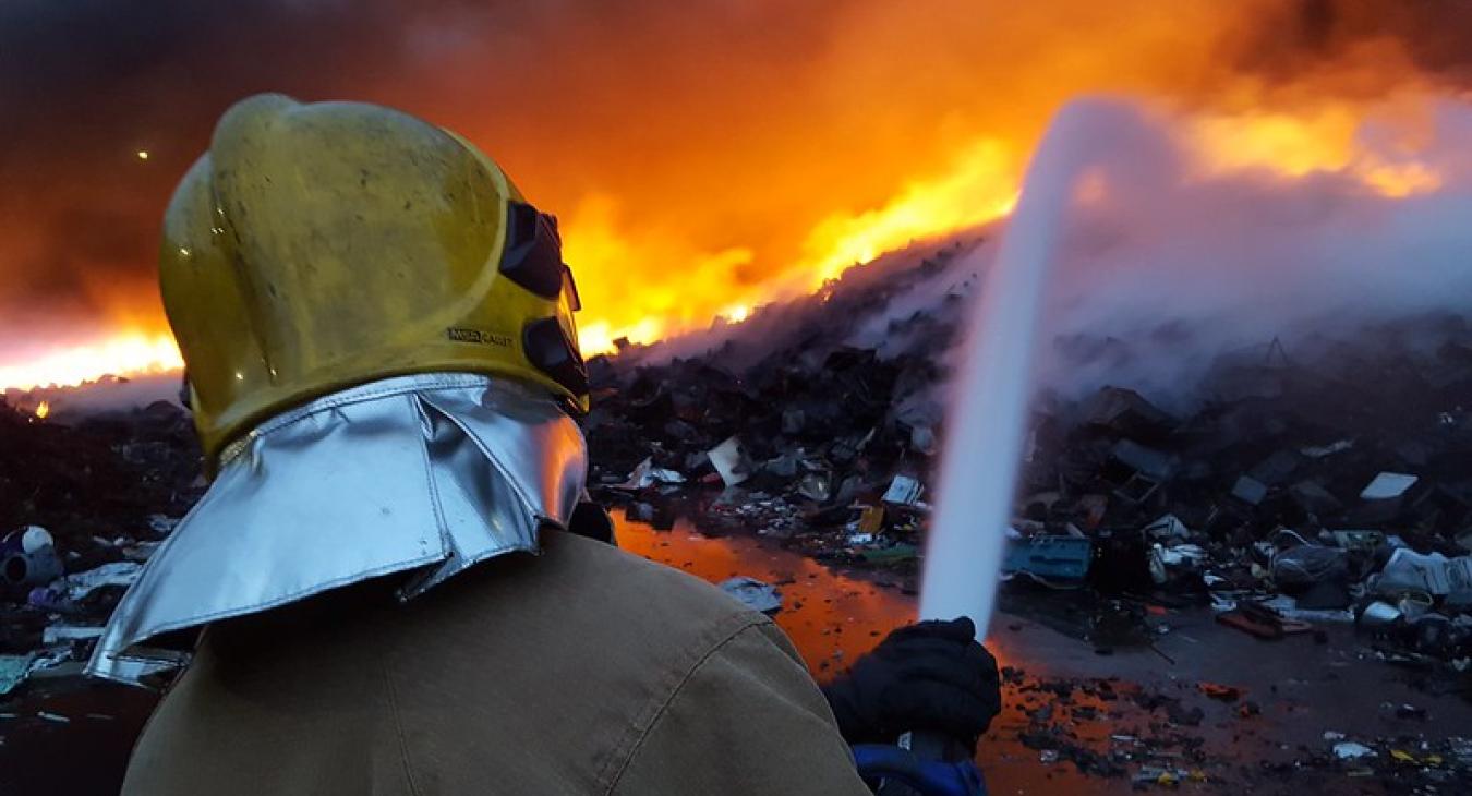 A helmetted firefighter shoots a big jet of water into a burning pile of recycling, which is glowing orange
