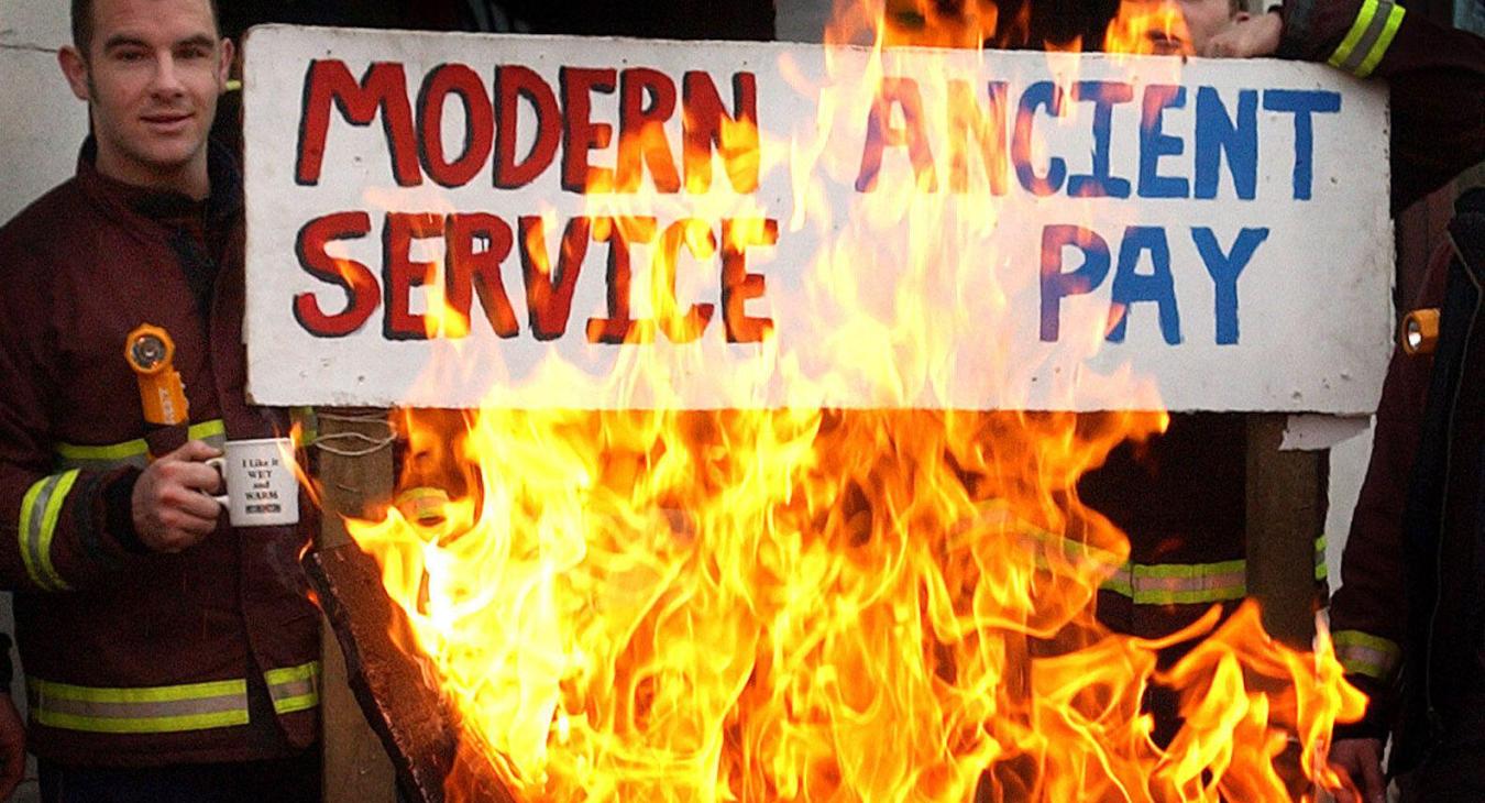 Firefighters behind a picket line brazier hold up a placard reading "modern service, ancient pay"