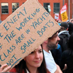 Woman holding a sign that reads "The enemy of the working class are in yachts not small boats"