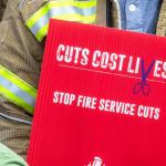 A firefighter holds a sign reading 'Cuts cost lives, stop fire service cuts'