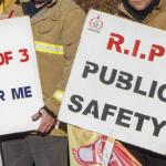 Two firefighters hold signs reading 'crews of three, not for me' and 'RIP public safety'