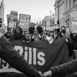 Black and white photo of a march outside Parliament with women holding signs saying cut warfare not welfare