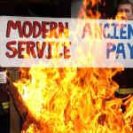 Firefighters behind a picket line brazier hold up a placard reading "modern service, ancient pay"