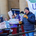 Tam Macfarlane wearing an FBU beanie speaks with a mic at a rally of striking phlebotomists